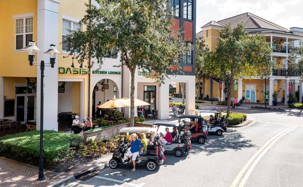 People sit in golf carts parked near a restaurant with outdoor seating under umbrellas on a sunny day in a lively, colorful urban area with trees and surrounding buildings. South Florida Business & Wealth