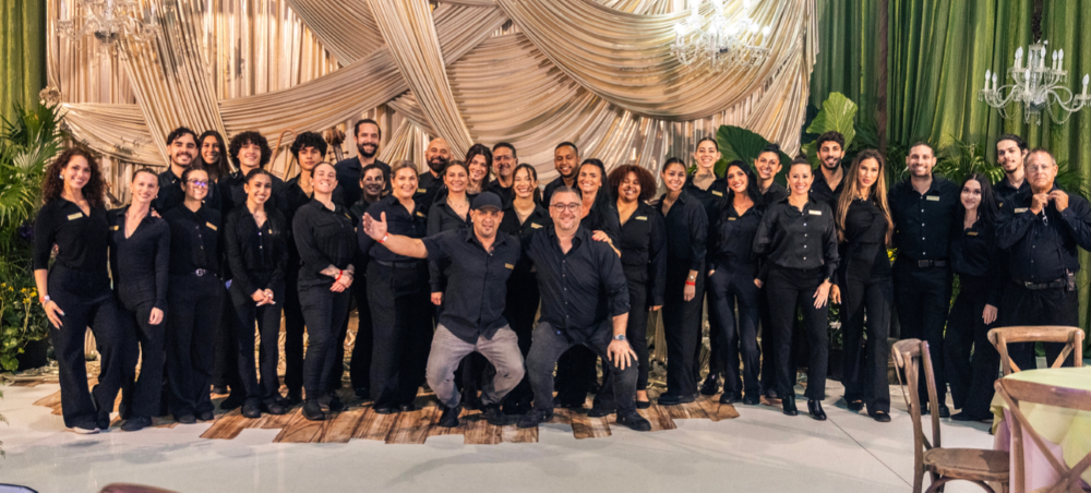 A large group of people dressed in black pose together indoors, smiling in front of decorative draped fabric and green curtains, under chandeliers. South Florida Business & Wealth