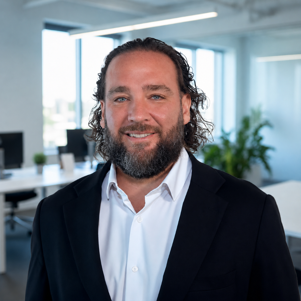 Smiling man with curly hair and beard in a suit jacket and white shirt stands in a modern, bright office with desks, computers, and large windows in the background. South Florida Business & Wealth