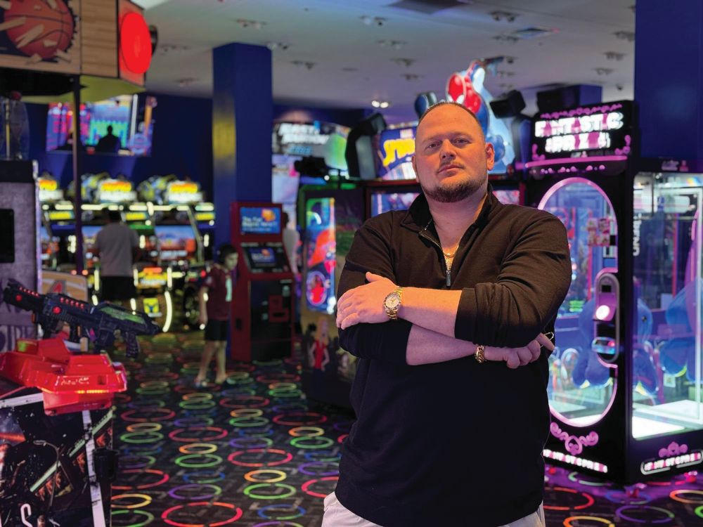 A man with short hair and a beard stands with arms crossed in a colorful arcade filled with games and neon lights. South Florida Business & Wealth