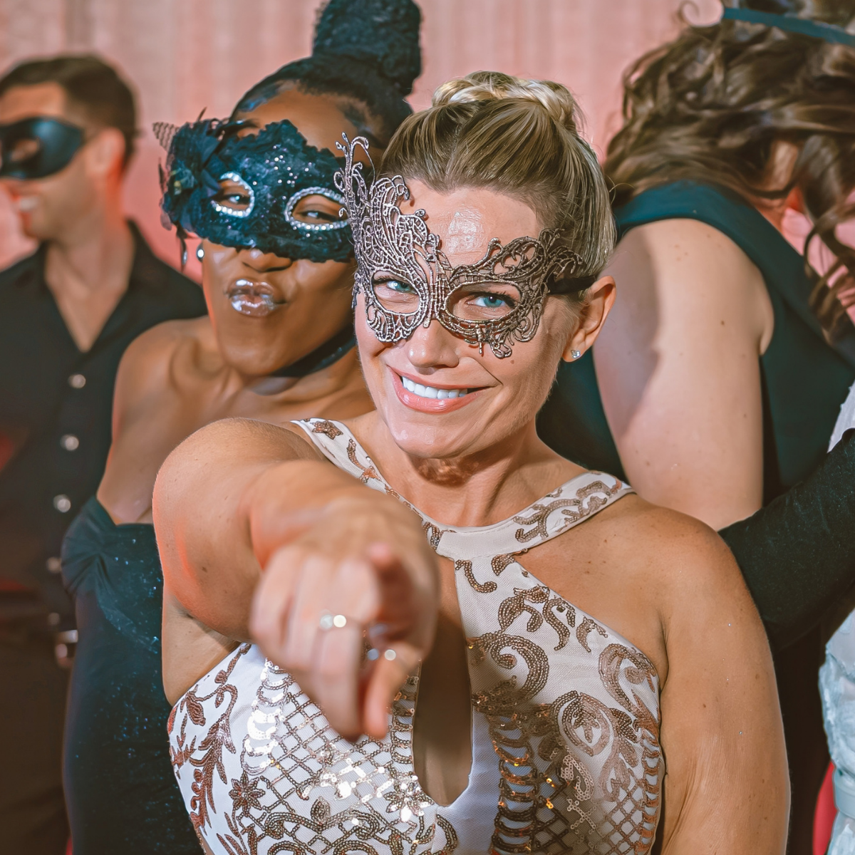 A woman in a fancy mask and dress smiles and points at the camera, surrounded by others in elegant attire and masks at a masquerade party. South Florida Business & Wealth
