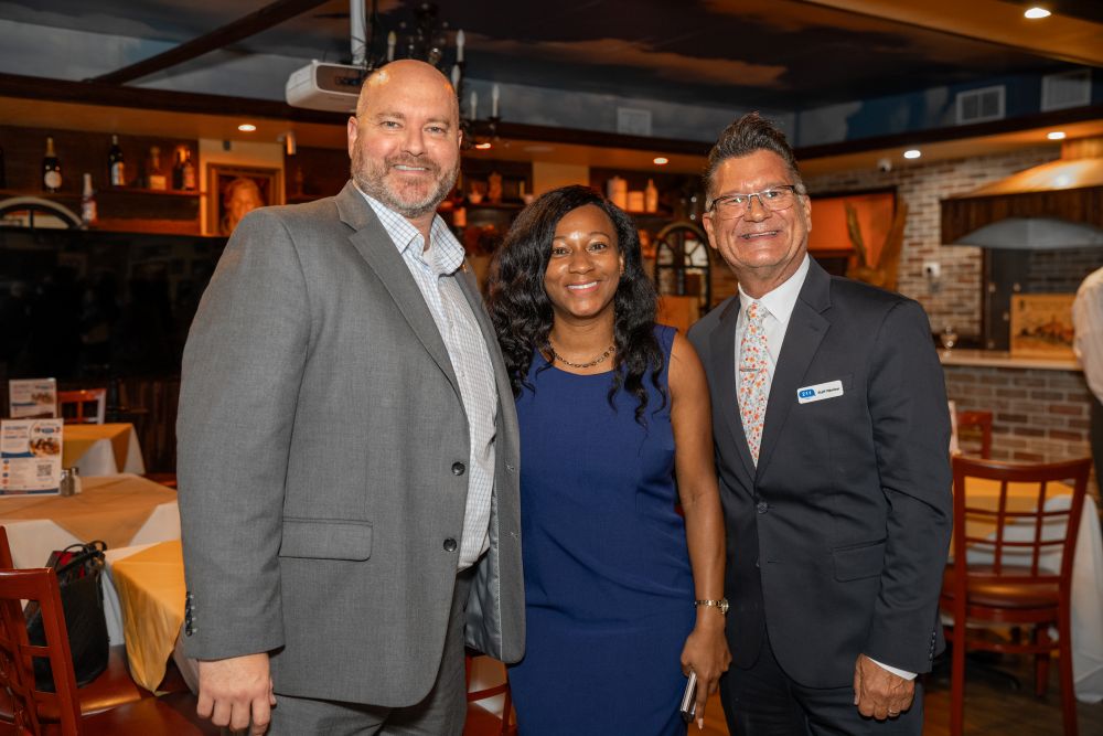 Three people in business attire stand smiling together at a restaurant, with tables, chairs, and shelves visible in the background. South Florida Business & Wealth