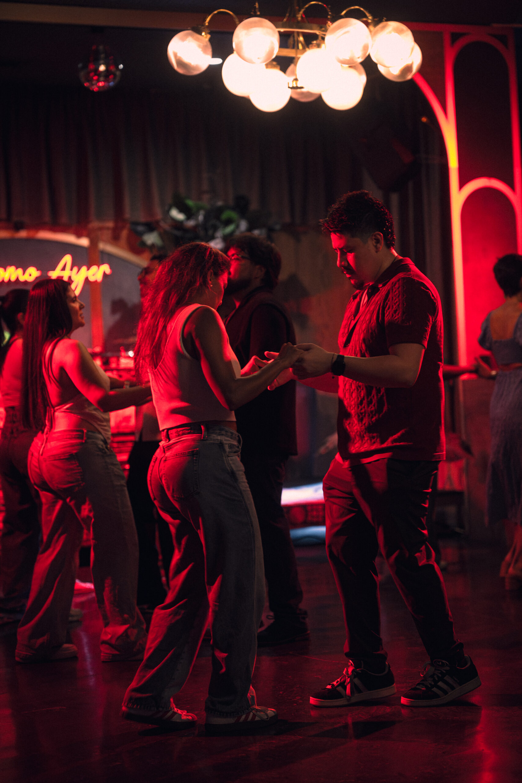 People dance in a dimly lit room with red lighting. A chandelier hangs overhead, and a neon sign glows in the background. Two people in the foreground face each other, holding hands on the dance floor. South Florida Business & Wealth