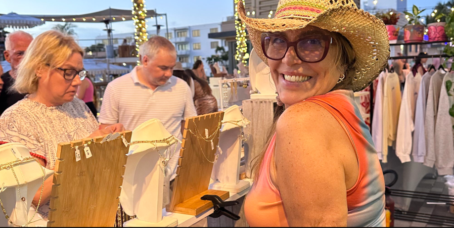 A woman in a hat and sunglasses smiles at a jewelry stand outdoors. Other shoppers browse nearby, and the area is decorated with string lights and clothing racks. South Florida Business & Wealth