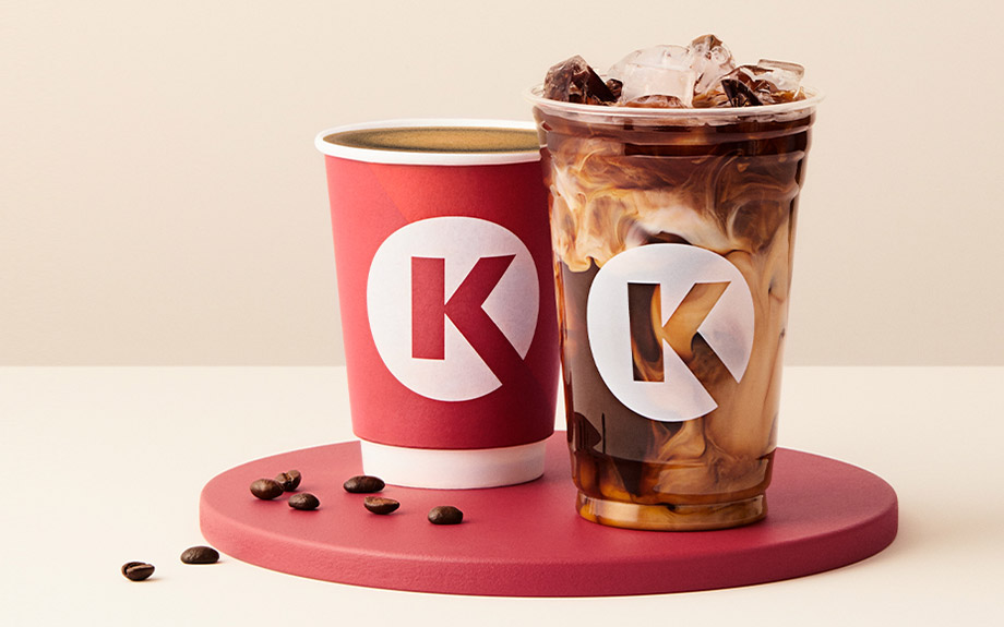 Two Circle K branded drinks: a hot coffee in a red paper cup and an iced coffee with milk swirls in a clear plastic cup, both on a red round platform with scattered coffee beans. South Florida Business & Wealth