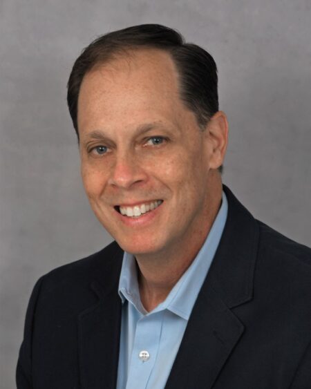 Smiling man with short brown hair wearing a dark blazer and light blue collared shirt, posing in front of a plain gray background. South Florida Business & Wealth