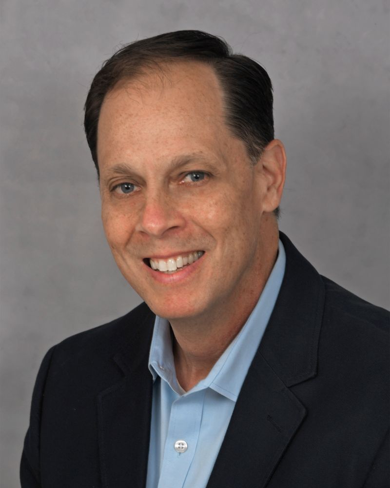 Smiling man with short brown hair wearing a dark blazer and light blue collared shirt, posing in front of a plain gray background. South Florida Business & Wealth