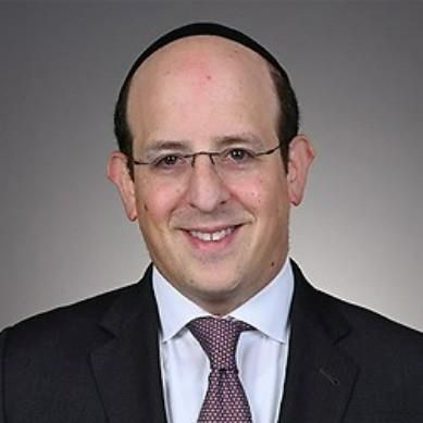 A man wearing glasses, a dark suit, white shirt, and patterned tie smiles at the camera against a plain gray background. South Florida Business & Wealth