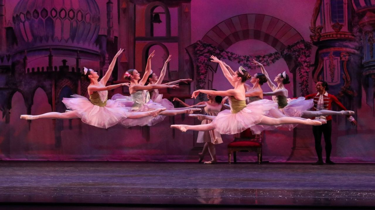Several ballet dancers in pink tutus perform a synchronized grand jeté leap on stage in front of a painted backdrop. South Florida Business & Wealth