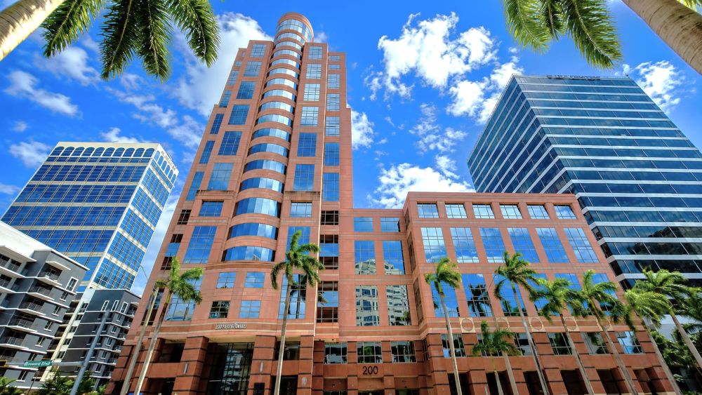 A tall, terracotta-colored office building with a curved glass facade, flanked by two modern glass-paned skyscrapers under a blue sky with palm trees in the foreground. South Florida Business & Wealth