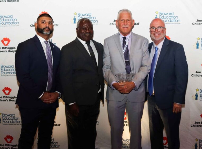 Four men in suits stand in a row in front of a step-and-repeat banner for the Broward Education Foundation, with the man second from the right holding a glass award. South Florida Business & Wealth