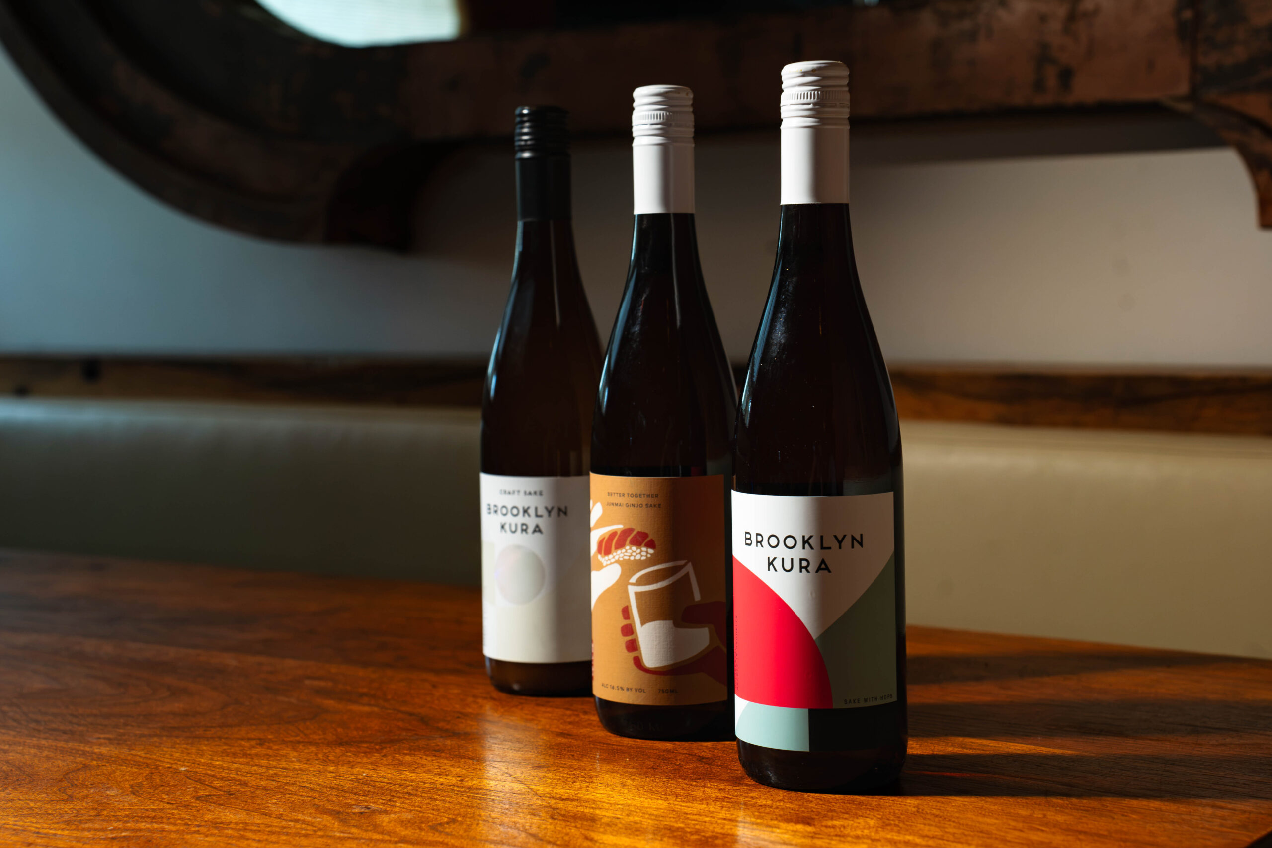 Three bottles of Brooklyn Kura sake with distinct colorful labels standing on a wooden table. South Florida Business & Wealth