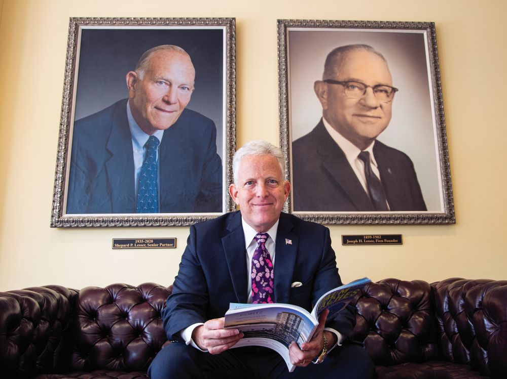 A man in a suit and butterfly tie sits on a leather sofa holding an open brochure, positioned between two framed portraits of men on the wall behind him. South Florida Business & Wealth
