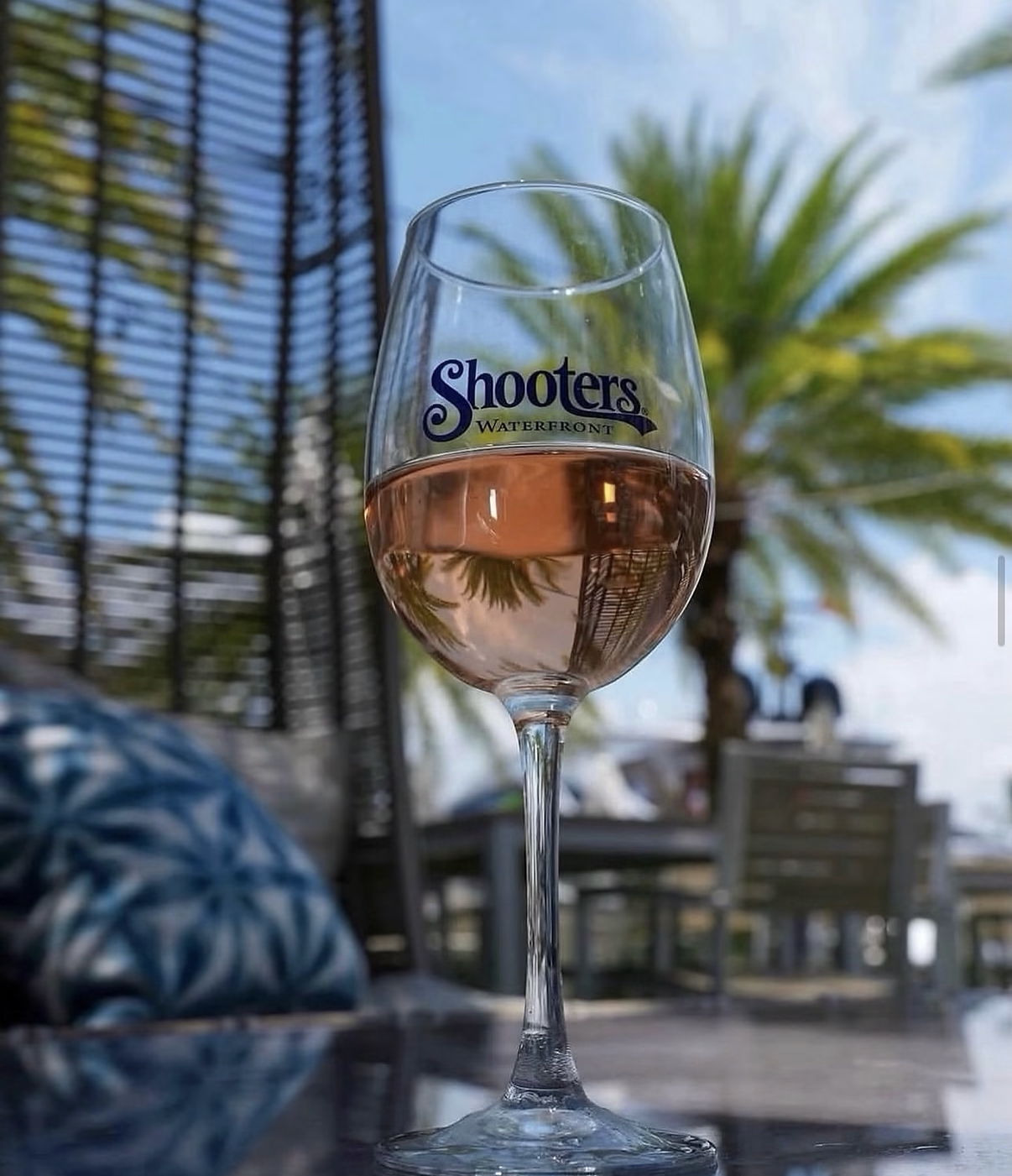 A glass of rosé wine branded with the “Shooters Waterfront” logo sits on a table outdoors with palm trees in the background. South Florida Business & Wealth