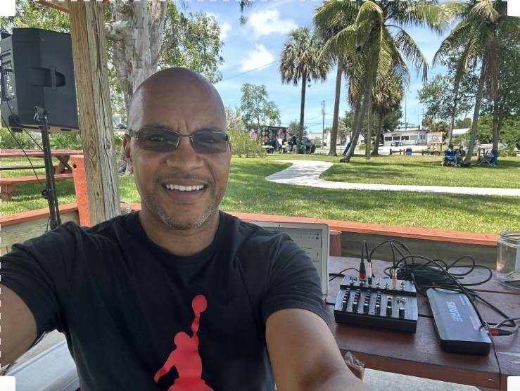 A smiling man wearing glasses and a dark t-shirt sits at an outdoor table with a small audio mixer and Shure equipment. South Florida Business & Wealth