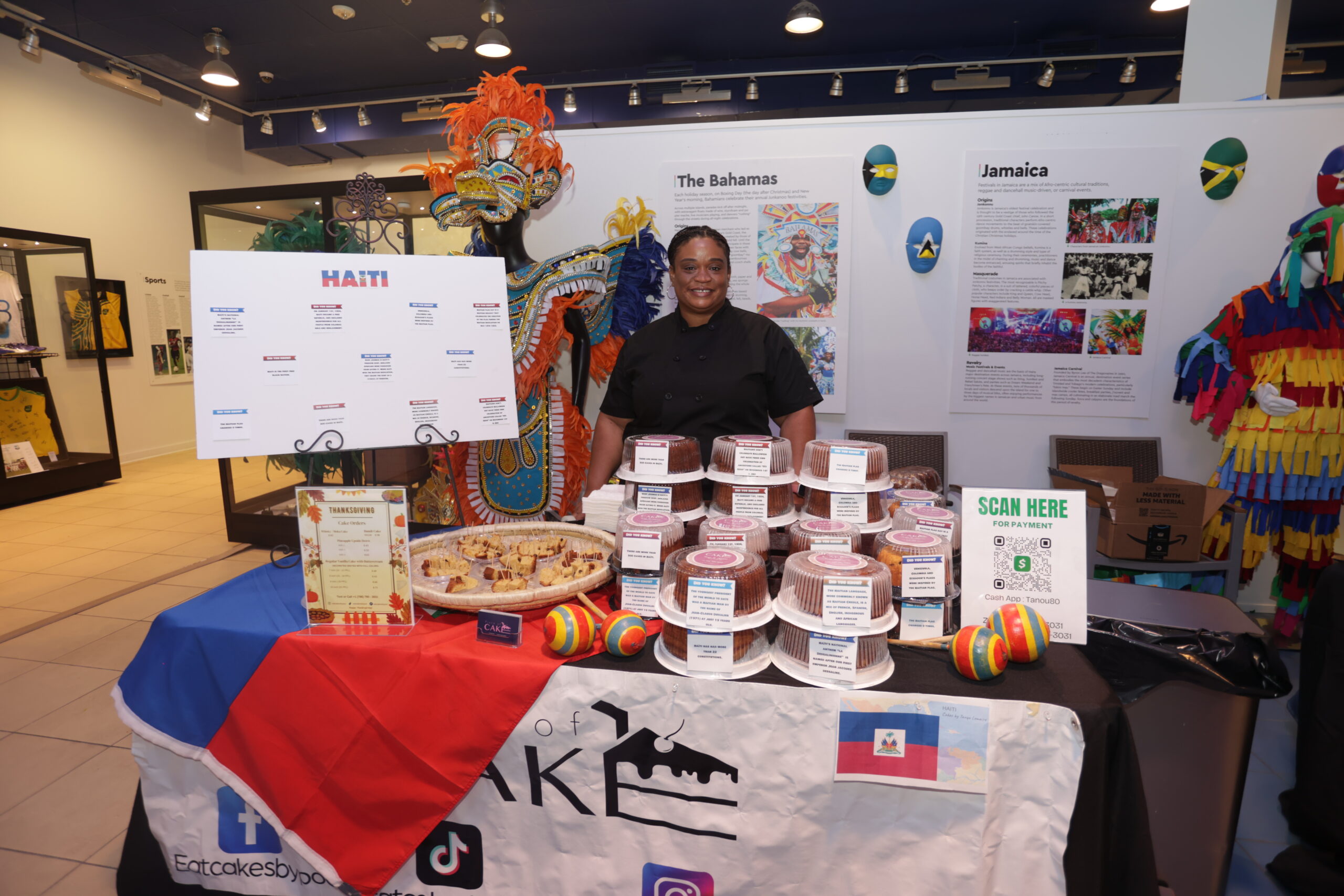 A woman stands behind a table displaying various cakes, a plate of pastries, and Haitian-themed decor, including a cultural display about The Bahamas and Jamaica in the background. South Florida Business & Wealth