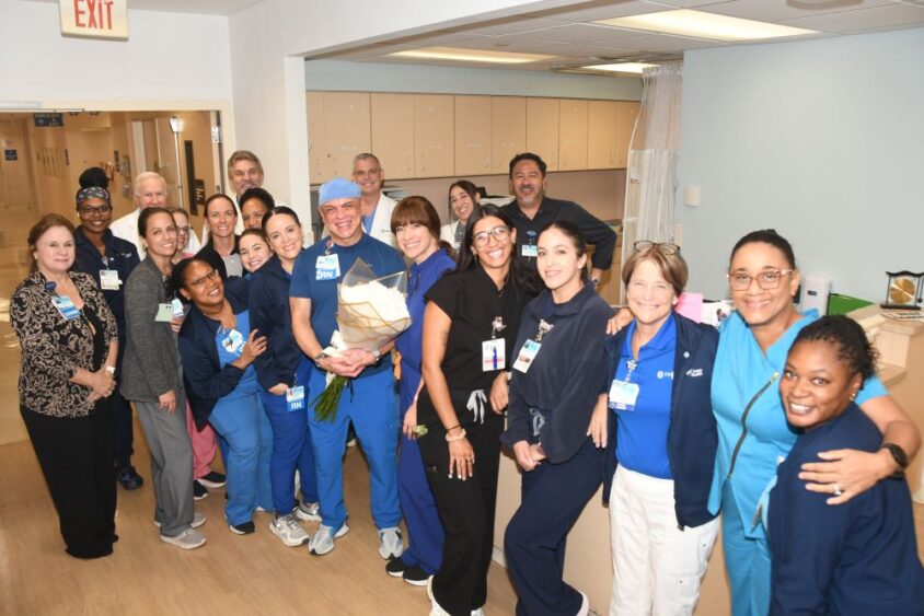 A large group of hospital staff posing together, with one nurse in the center holding a large bouquet of white flowers. South Florida Business & Wealth
