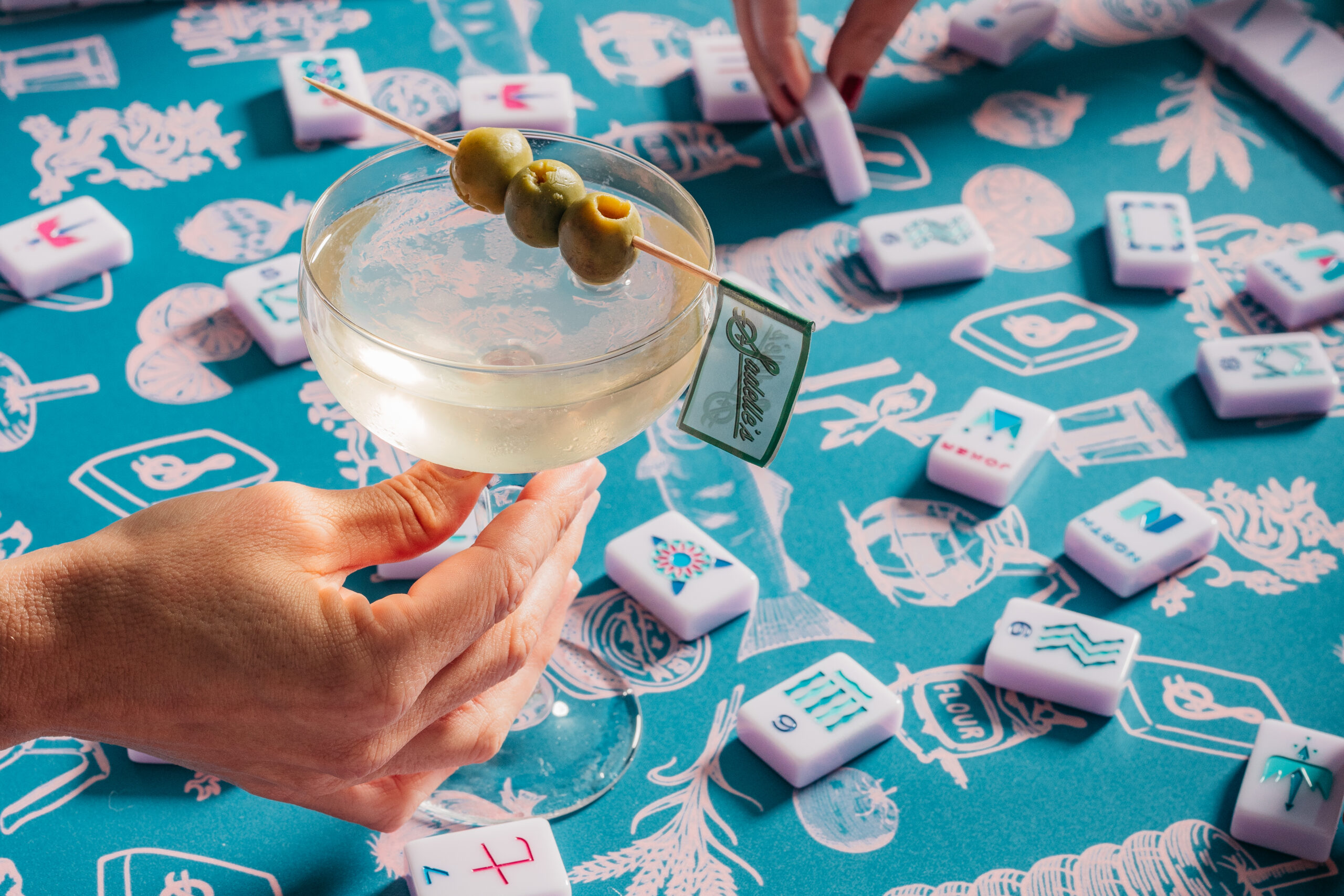 A hand holds a martini garnished with three olives on a pick, positioned over a table with a patterned blue tablecloth and scattered mahjong tiles. South Florida Business & Wealth