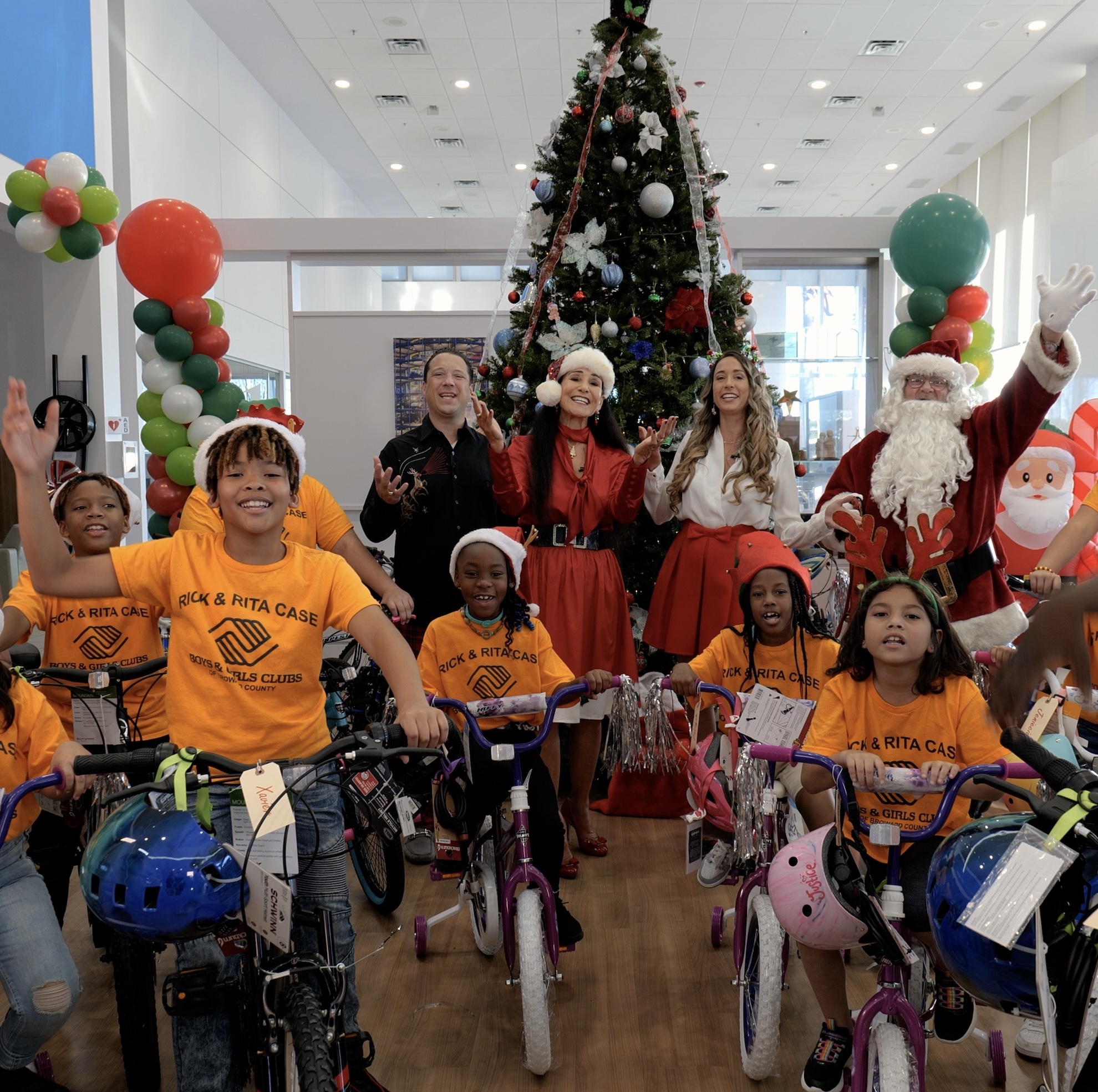 A group of children in yellow Boys & Girls Club t-shirts pose with new bicycles alongside adults, including Santa Claus, in front of a decorated Christmas tree. South Florida Business & Wealth