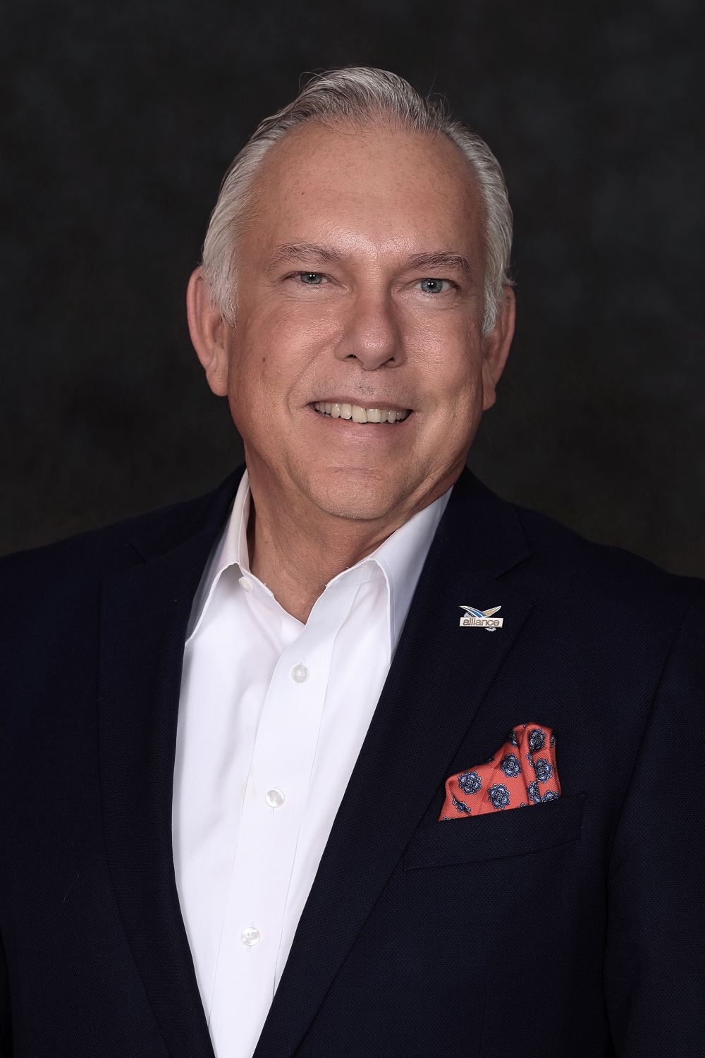 A middle-aged man with gray hair wearing a dark blue suit jacket, white dress shirt, and a patterned red pocket square with an Alliance lapel pin. South Florida Business & Wealth