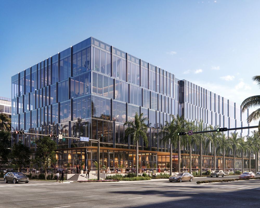 A modern multi-story office building with a glass facade, ground-floor retail spaces, and surrounding palm trees on a city street corner. South Florida Business & Wealth