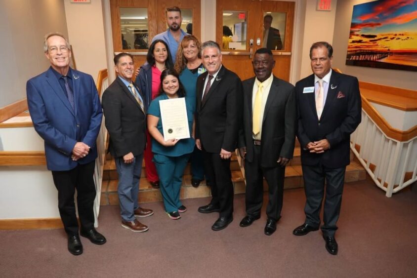 A group of seven people posing for a photo, with the woman in the center holding a framed certificate. South Florida Business & Wealth