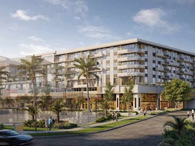 A multi-story apartment complex with ground-floor retail shops, palm trees, and a paved walkway along a waterfront. South Florida Business & Wealth